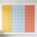Calendrier mural familial réaliste avec une grille mensuelle vierge organisée en trois colonnes colorées jaune, bleue et corail, dans une cuisine lumineuse.